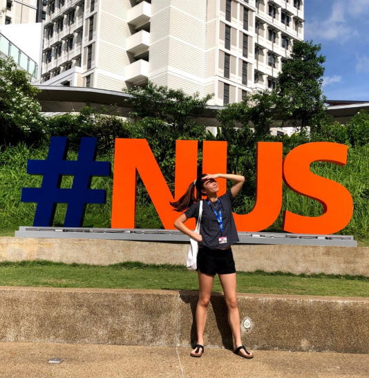 Aliya smiling in front of a #NUS sign