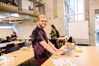 A student smiling at the camera while using his laptop in the Engineering Design Centre