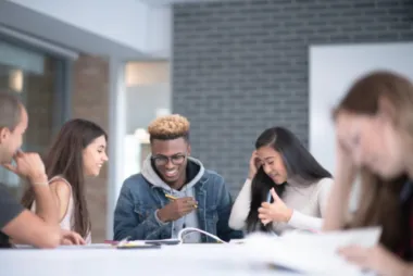 a group of five students sitting around a table, one reading book alone while the others are discussing ideas