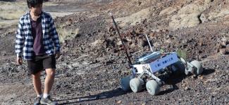 Rover team member walking alongside prototype on a rocky hillsie